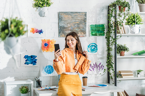 happy stylish girl using smartphone in room decorated with green plants and colorful paintings on white wall