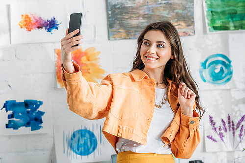 cheerful young woman taking selfie while standing near white wall with colorful paintings