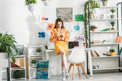 attractive young woman using smartphone in room decorated with green plants and paintings on wall