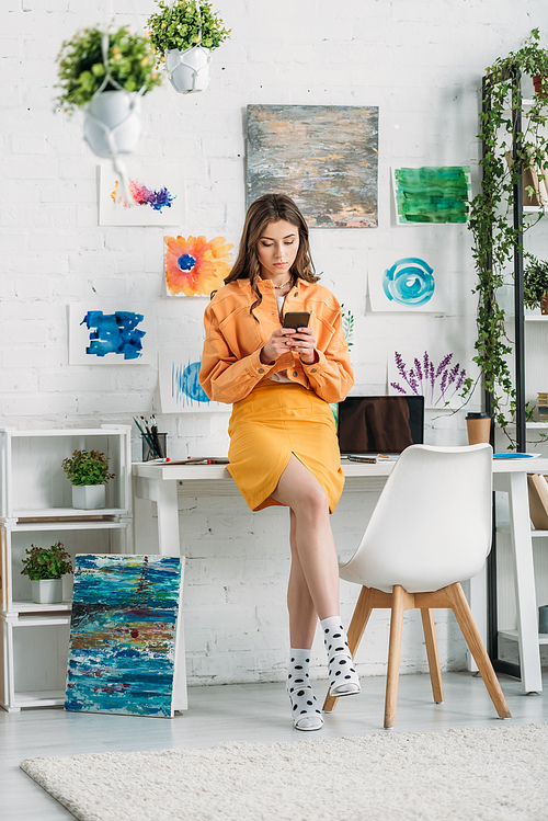 stylish young woman using smartphone in spacious room decorated with green plants and colorful paintings on wall