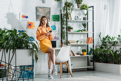 stylish grl using smartphone in spacious room decorated with green plants and colorful paintings on wall
