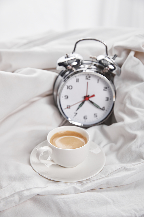 coffee in white cup on saucer near silver alarm clock in white bedding