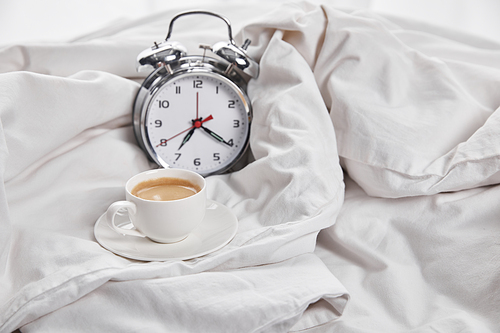 coffee in white cup on saucer near silver alarm clock in white bed