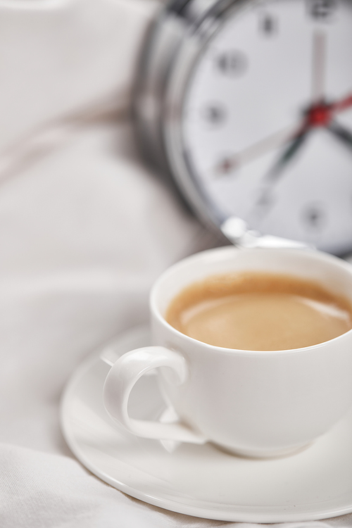 selective focus of coffee in white cup on saucer with silver alarm clock in bed on background