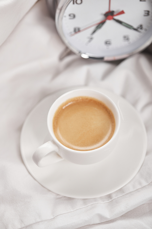 top view of coffee in white cup on saucer near silver alarm clock on bedding