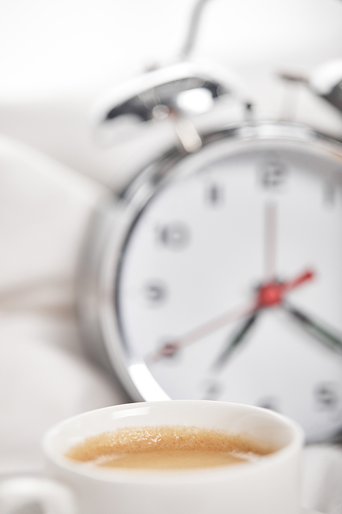 selective focus of coffee in white cup with silver alarm clock in bed on background
