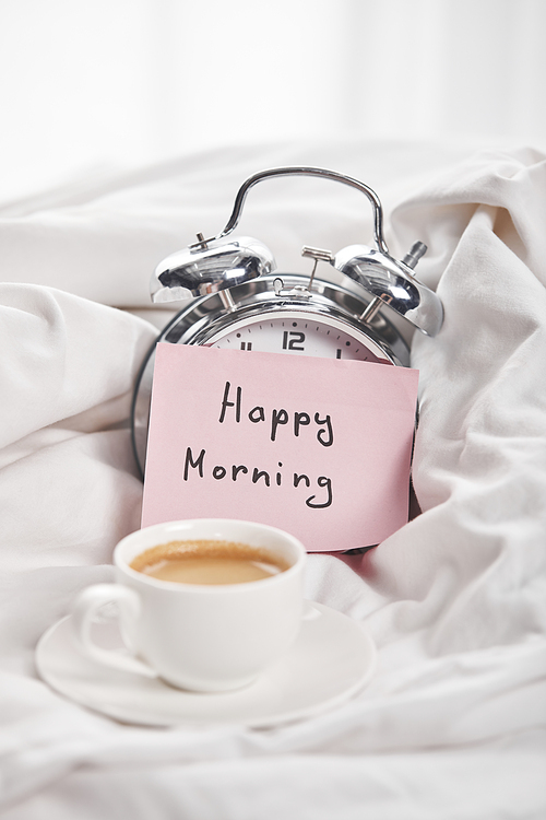coffee in white cup on saucer near silver alarm clock with happy morning lettering on sticky note in bed