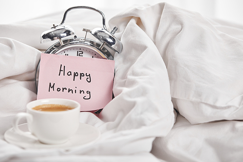 coffee in white cup near silver alarm clock with happy morning lettering on sticky note in bed