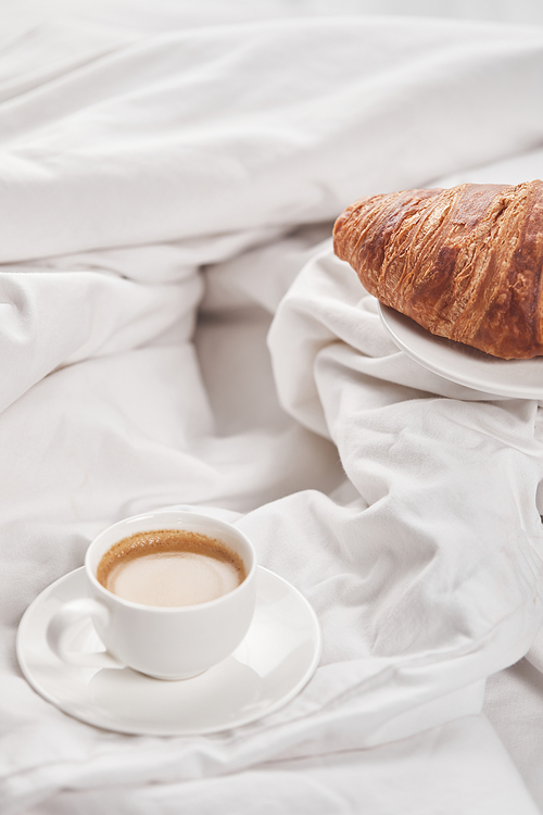 fresh tasty croissant on plate near coffee in white cup on saucer in bed