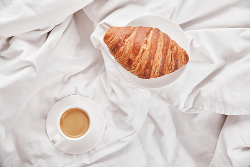 top view of fresh croissant on plate near coffee in white cup on saucer in bed