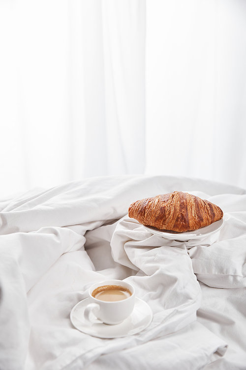 fresh croissant on plate near coffee in white cup on saucer in bed