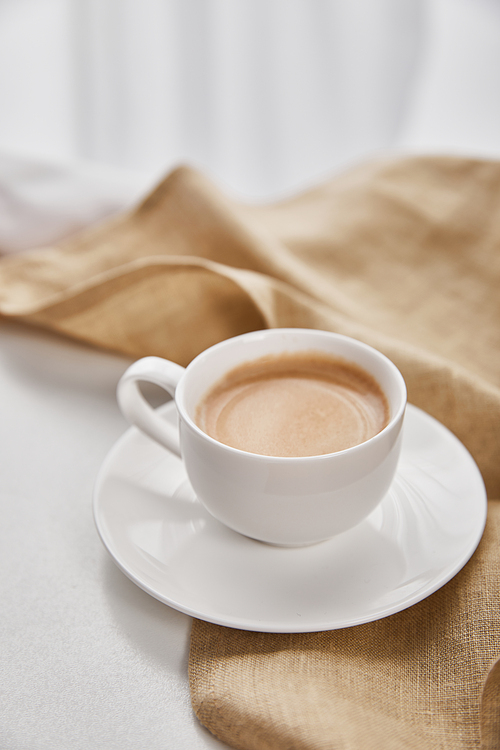close up view of coffee in white cup on saucer near beige napkin