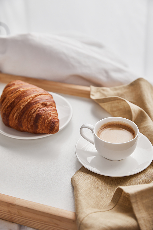 fresh croissant on plate with coffee in cup on wooden tray in bed at morning