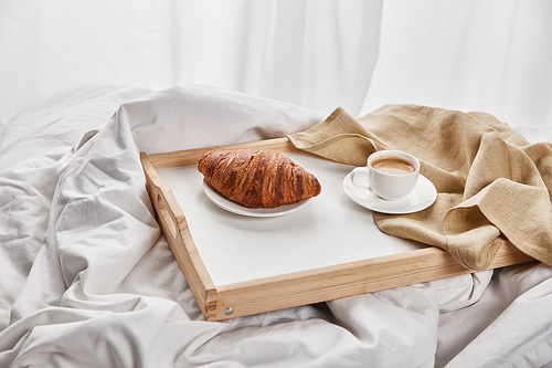 fresh croissant with coffee and napkin on wooden tray in bed at morning