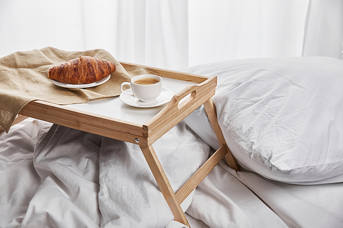 fresh croissant with coffee on wooden tray in bed with pillow at morning