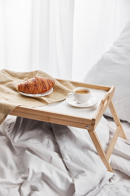 coffee and croissant served on wooden tray on white bed with pillow