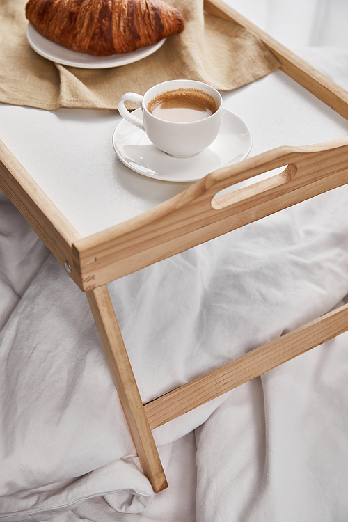 close up view of coffee and croissant served on wooden tray on white bed