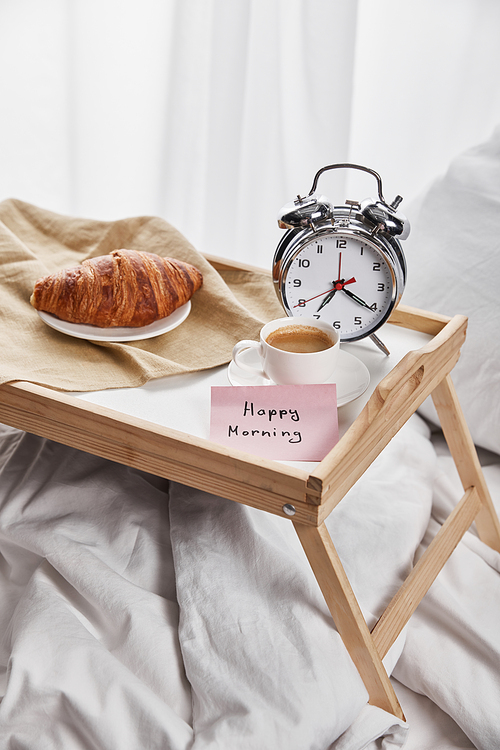 silver alarm clock, sticky note with happy morning lettering, coffee and croissant on wooden tray on white bedding