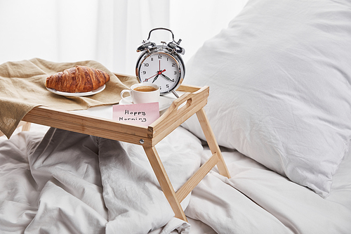 alarm clock, sticky note with happy morning lettering, coffee and croissant on wooden tray on white bedding