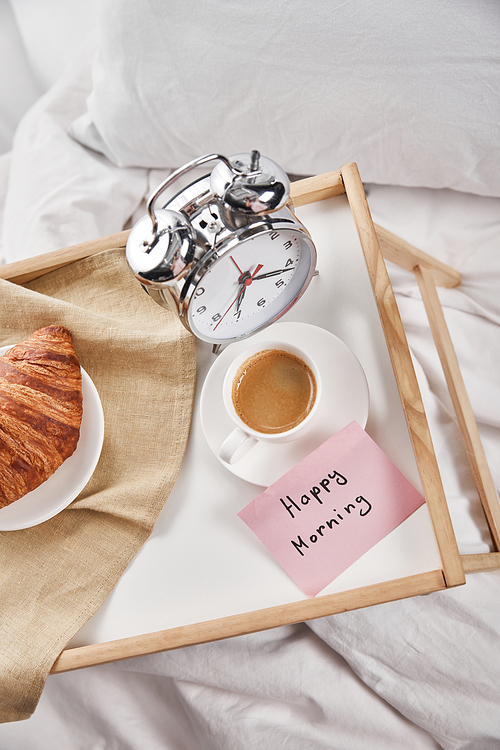 top view of alarm clock, sticky note with happy morning lettering, coffee and croissant on wooden tray on white bedding