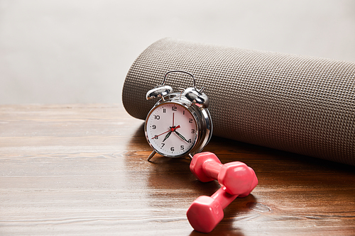 silver alarm clock near dumbbells and fitness mat on wooden table isolated on grey