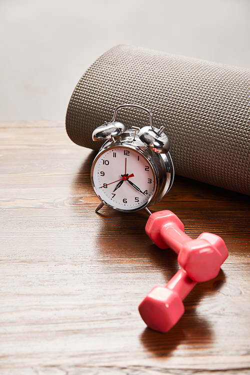 silver alarm clock near pink dumbbells and fitness mat on wooden table isolated on grey