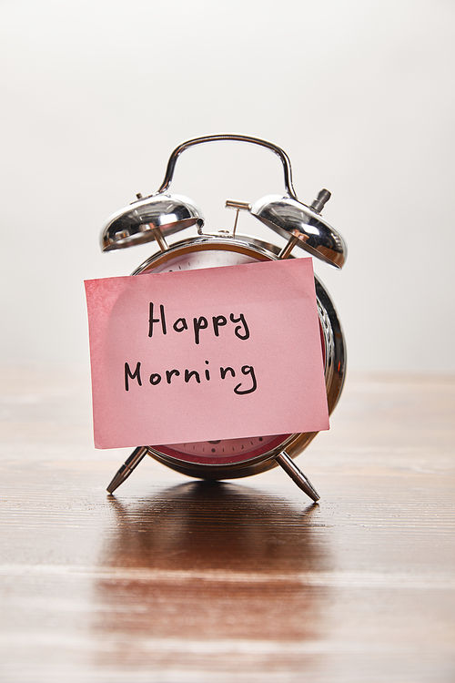 silver alarm clock with happy morning lettering on pink sticky note on wooden table isolated on grey