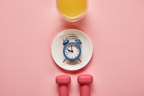 top view of dumbbells and plate with toy alarm clock near glass with orange juice on pink background