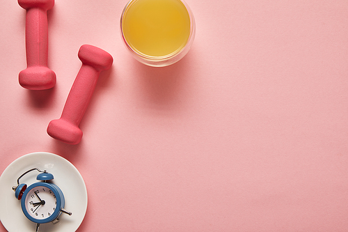 top view of orange juice, pink dumbbells and toy alarm clock on plate on pink background