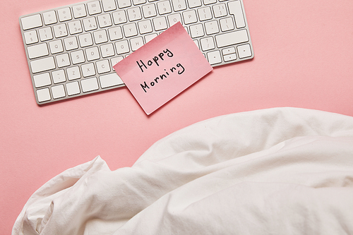 top view of blanket, computer keyboard and pink sticky note with happy morning lettering on pink background