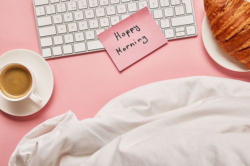 top view of blanket, computer keyboard and pink sticky note with happy morning lettering near croissant and coffee on pink background