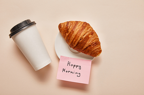 top view of coffee to go, croissant and sticky note with happy morning lettering on beige background