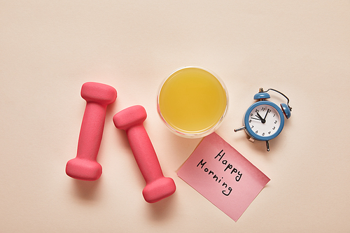 top view of sticky note with happy morning lettering, pink dumbbells, orange juice and small alarm clock on beige background
