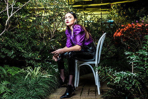 Attractive woman in purple blouse and leather pants sitting on chair and holding wine glass in orangery