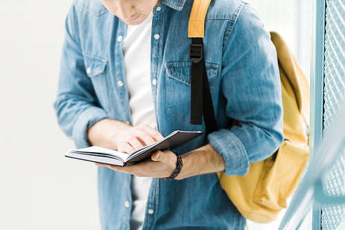 partial view of student in denim shirt with yellow backpack reading notebook