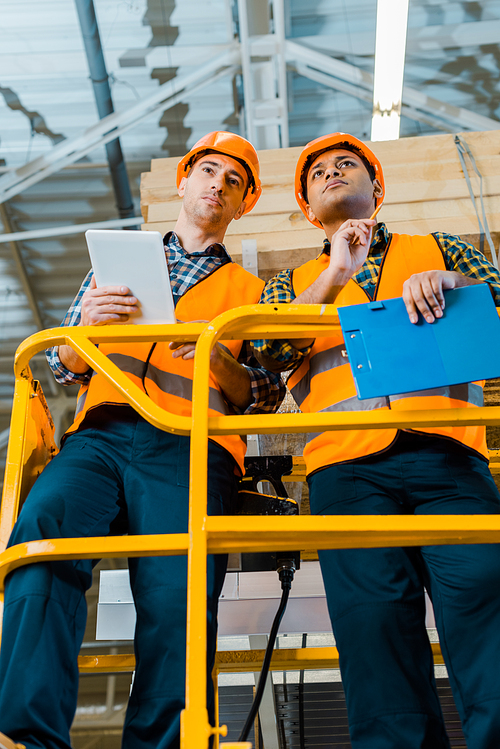 concentrated multicultural warehouse workers with digital tablet and clipboard standing on scissor lift