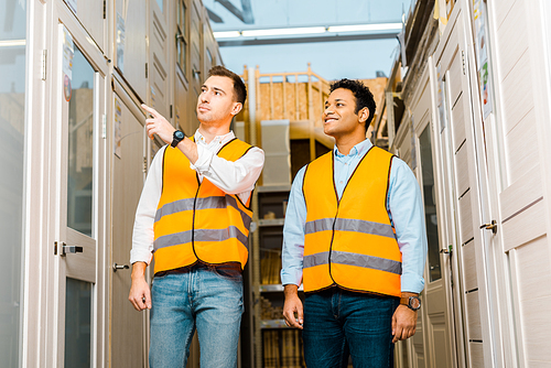 handsome warehouse worker pointing with finger at doors near smiling indian coworker