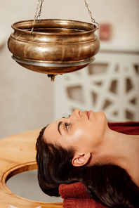 relaxed young woman lying under shirodhara vessel during ayurvedic procedure