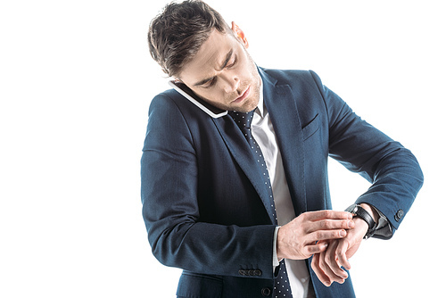 handsome, serious businessman talking on smartphone and looking at watch isolated on white