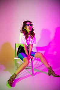 attractive young woman in sunglasses sitting on chair and showing tongue on purple with gradient