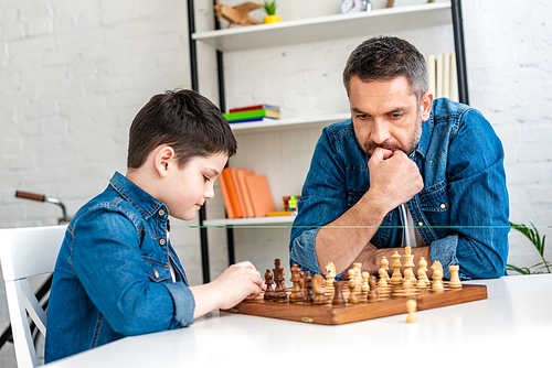 pensive father and son in denim playing chess while sitting at table at home