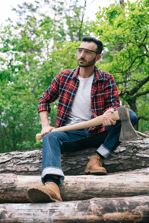 thoughtful lumberman holding ax while sitting on tree trunks in forest and looking away