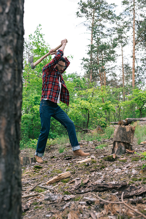 selective focus of lumberjack in plaid shirt and denim jeans cutting wood with ax in forest