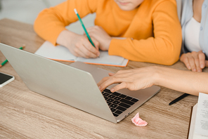 cropped view of woman using laptop and boy writing in copy book while doing schoolwork at home