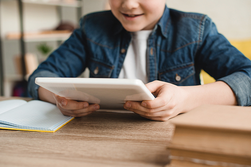 partial view of smiling boy using digital tablet while sitting at desk with books at home