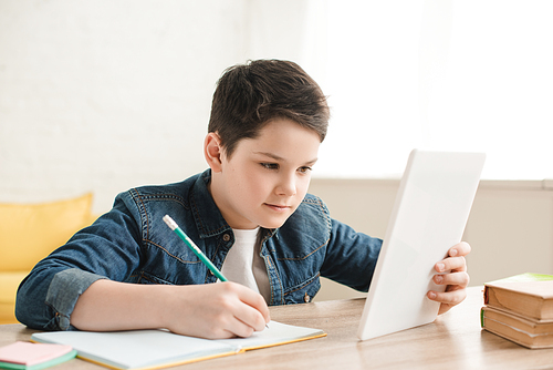 concentrated boy writing in notebook and using digital table while doing schoolwork at home