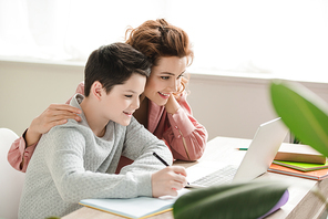 happy mother hugging adorable son while doing schoolwork together at home