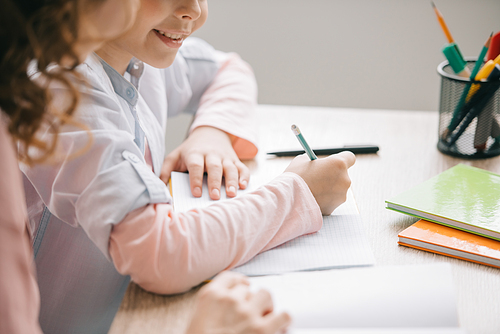 cropped view of mother and daughter sitting at table and doing schoolwork together at home