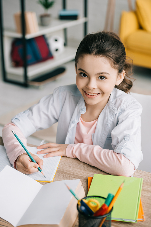 cheerful schoolkid writing in notebook and smiling at camera while doing homework