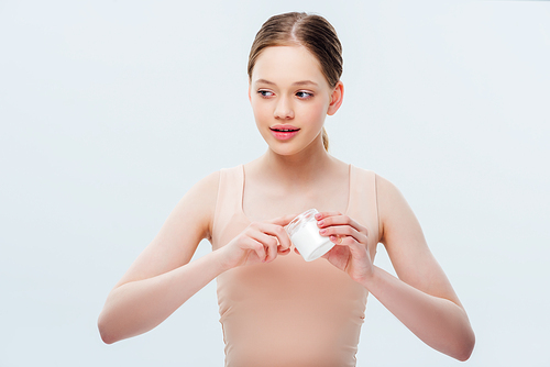 smiling teenage girl applying cosmetic cream on fingers and looking away isolated on grey